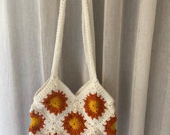 May include: A white crochet bag with a floral pattern in orange and yellow. The bag has two white straps and is hanging against a white curtain. The bag is handmade and has a unique, textured appearance.