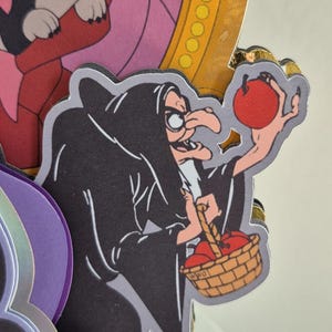 May include: A layered paper cutout featuring a cartoon witch in a black cloak holding a red apple and a basket of apples. The witch has a long nose and is wearing glasses. The background includes other cartoon characters and a gold-colored frame.