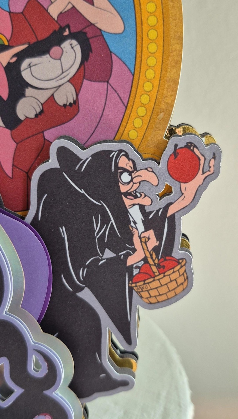 May include: A layered paper cutout featuring a cartoon witch in a black cloak holding a red apple and a basket of apples. The witch has a long nose and is wearing glasses. The background includes other cartoon characters and a gold-colored frame.