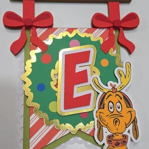 May include: A festive banner with a red letter "E" on a green and red background, accented with gold trim and a cartoon character. The banner is adorned with red bows and a brown wooden bar.