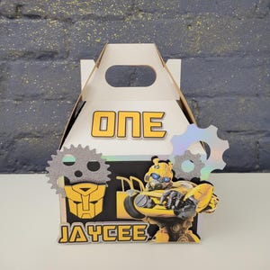 May include: A white gable box with the word "ONE" in yellow letters. The box is decorated with a yellow and black Transformer character and the name "JAYCEE". Two silver gear-shaped embellishments are on the sides.