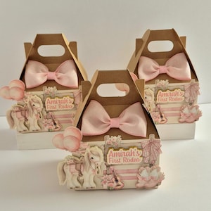 Puede incluir: Tres cajas de regalo de cartón marrón con lazos rosas y adornos temáticos de rodeo. Cada caja presenta un diseño rosa y blanco con el texto "Amirah's First Rodeo", un caballo de dibujos animados y una guitarra.