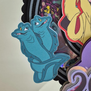May include: A layered paper craft featuring cartoon illustrations of Disney villains. The central focus is on two teal-colored sea serpent characters. Other characters include Cruella de Vil and Ursula.