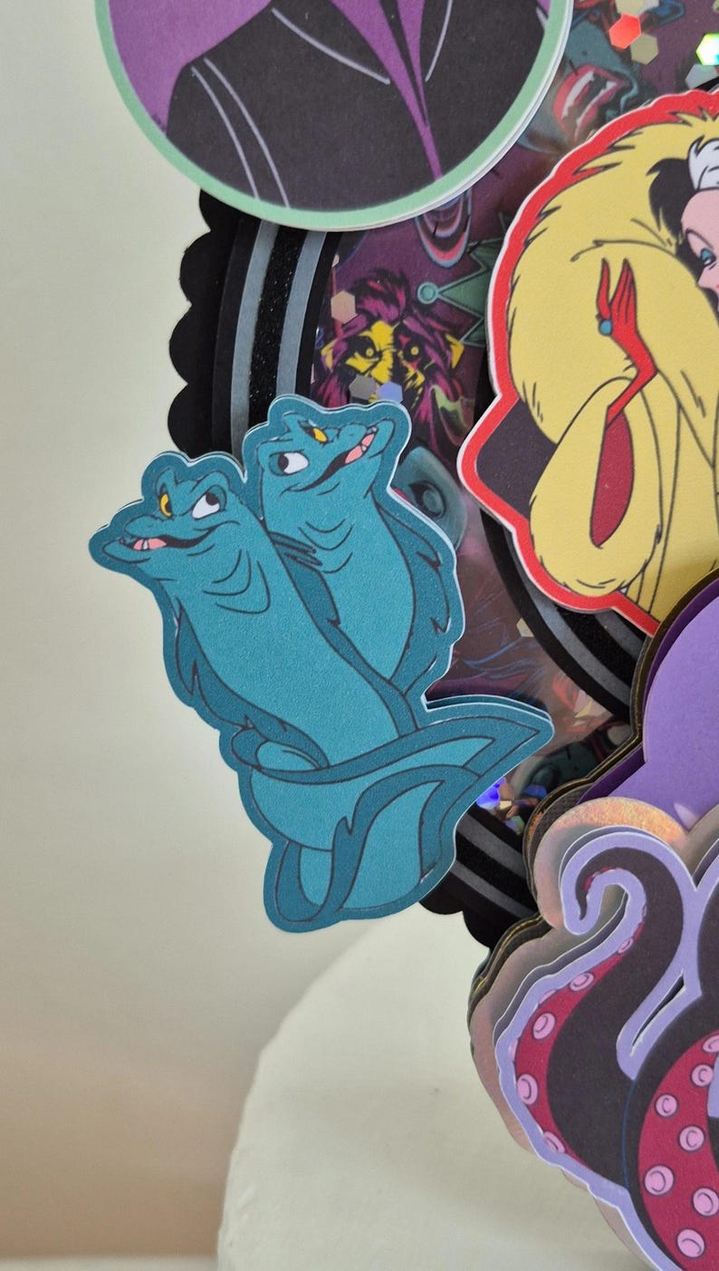 May include: A layered paper craft featuring cartoon illustrations of Disney villains. The central focus is on two teal-colored sea serpent characters. Other characters include Cruella de Vil and Ursula.