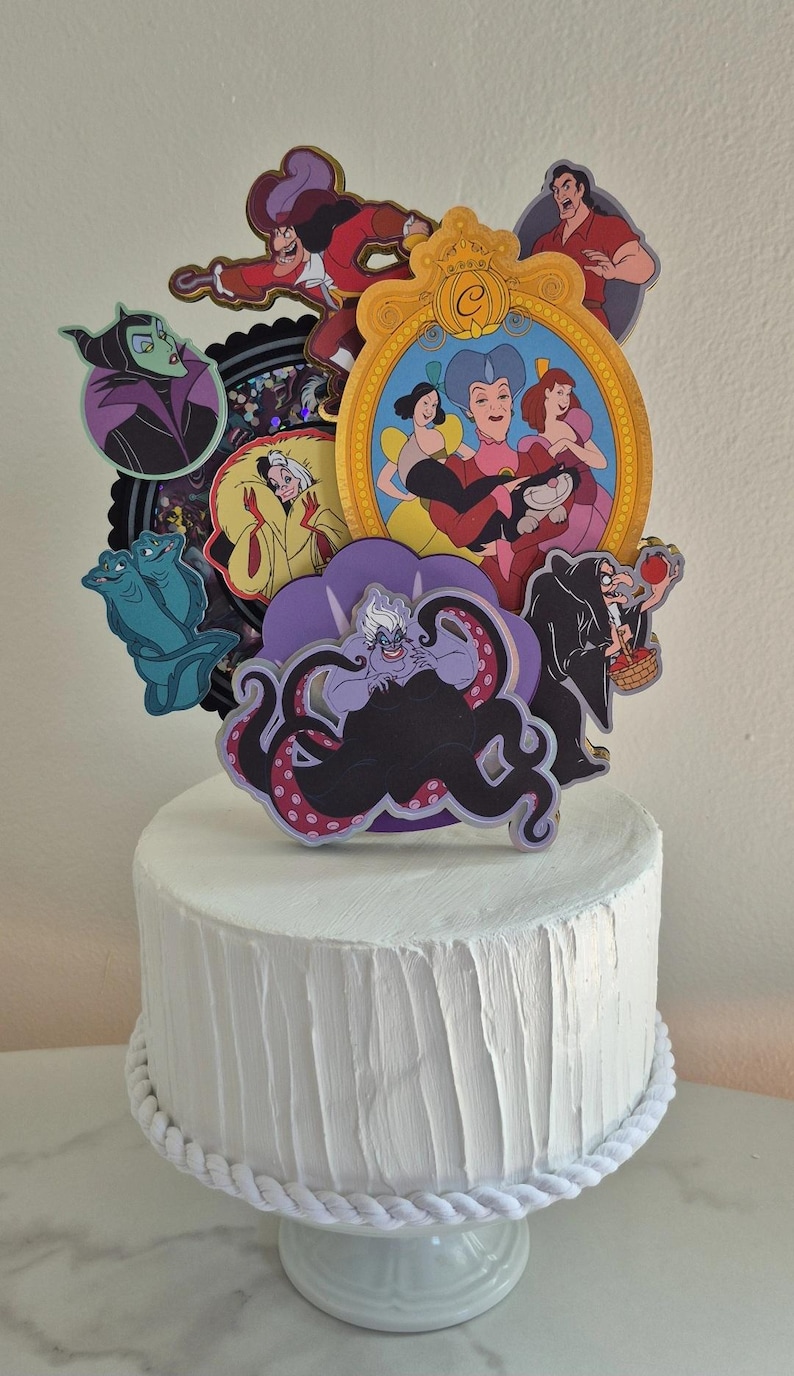 May include: A white cake with a textured surface, adorned with colorful cutouts of Disney villains. The cake is decorated with a white rope trim and sits on a white pedestal. The cutouts include Ursula, Maleficent, and Captain Hook.