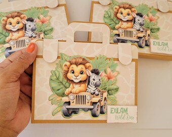 May include: Safari-themed gift bags with a lion and zebra in a jeep design. The bags have a handle and are decorated with tropical leaves and flowers. The text "EILAM Wild One" is printed on a small label. The bags are beige and brown.