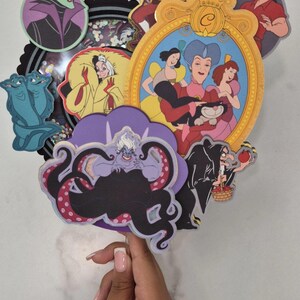 May include: A collection of colorful cartoon character cutouts, including Ursula, Maleficent, and Captain Hook. The characters are arranged in a layered design, with a gold-framed image of Cinderella and her stepfamily. The overall theme is Disney villains.
