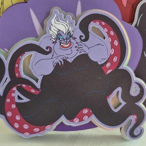 May include: A purple Ursula the Sea Witch character cutout with white hair, red lipstick, and black tentacles with pink spots. The cutout is surrounded by other cartoon character cutouts.