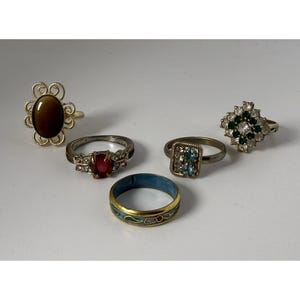 May include: A collection of five rings: one with a brown oval stone in a filigree setting, one with a red oval stone and clear accents, one with a rectangular blue stone cluster, one with a green and clear stone cluster, and a gold band with blue enamel.