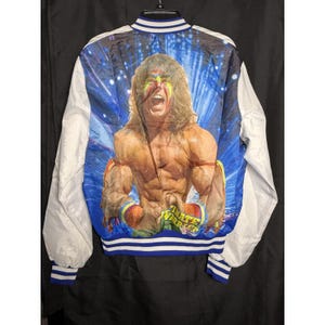 Könnte beinhalten: Eine weiß-blaue Bomberjacke mit einem großen, farbenfrohen Grafikdruck auf der Rückseite, der einen muskulösen Wrestler mit langen Haaren und Gesichtsbemalung zeigt. Der Text ULTIMATE WARRIOR steht auf dem Armband des Wrestlers. Die Jacke hat gestreifte Bündchen.