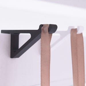 Op de afbeelding: Een zwarte, geometrische plankbeugel met een driehoekige steun, gemonteerd op een wit oppervlak. Een bruin lint hangt aan de beugel. Het ontwerp is minimalistisch en modern, geschikt voor interieurdecoratie.