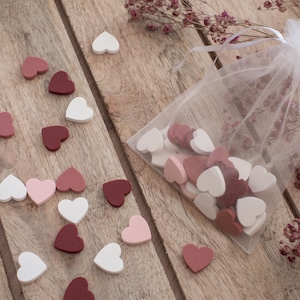 Può includere: Decorazioni a forma di cuore in legno bianco, rosa e bordeaux sparse su una superficie di legno. È presente anche un sacchetto trasparente pieno di cuori simili. Fiori secchi aggiungono un tocco delicato.