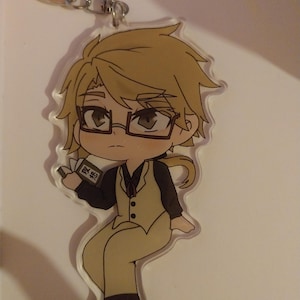 Peut inclure: Un porte-clés en acrylique transparent représentant un personnage de dessin animé aux cheveux blonds, portant des lunettes et un costume beige. Le personnage tient un petit livre avec du texte japonais. Le porte-clés a un fermoir en métal argenté.