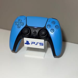 May include: A light blue PlayStation 5 controller with black accents, displayed on a white stand. The controller features the standard button layout and a touchpad. The stand displays the PS5 logo.