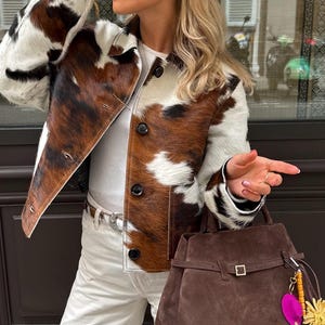 Puede incluir: Una chaqueta con estampado de piel de vaca con un bolso de mano de ante marrón. La chaqueta tiene un estampado marrón, negro y blanco. El bolso tiene una hebilla dorada y un adorno rosa y amarillo. La persona lleva pantalones blancos y gafas de sol.
