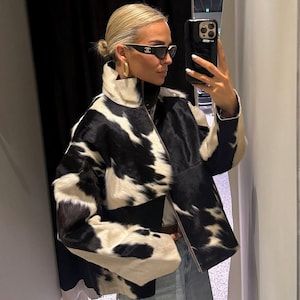 Pode incluir: Casaco de pele de vaca preto e branco com gola alta e fecho de correr. O casaco tem um padrão de manchas único e é combinado com jeans de ganga clara. A pessoa está a usar óculos de sol pretos com um logótipo branco.