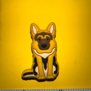 May include: A yellow, black, and brown German Shepherd dog charm. The charm is sitting with its legs together and tail curled. The dog has a white chest and black accents. The background is yellow.
