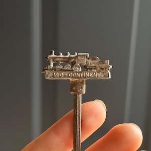 May include: A vintage silver spoon with a train-shaped top, featuring the text "MID-CONTINENT". The spoon's bowl has engraved text. The spoon is held in a hand against a gray background. A collectible souvenir.