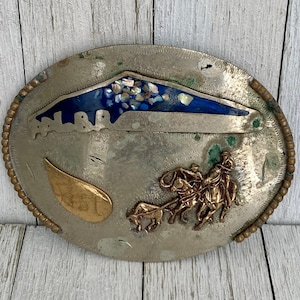 Peut inclure: Boucle de ceinture en argent avec une incrustation bleue et blanche représentant une scène de montagne. La boucle présente également un motif doré d'un cow-boy à cheval tirant un chariot.