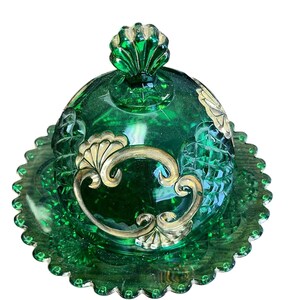 Puede incluir: Un mantequillero de vidrio verde con un diseño pintado en oro. El plato tiene un borde festoneado y una tapa abovedada con un pomo.
