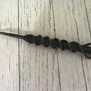 May include: A hand-forged, dark metal hook tool with a twisted handle design. The hook curves at the end, and the handle features a spiral pattern. The tool has a loop at the end of the handle.