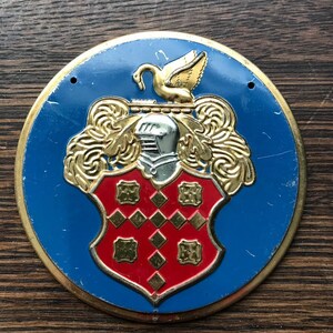 May include: A round, blue and gold metal emblem with a red shield featuring a knight's helmet, a swan, and a gold design.