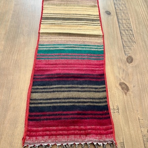 May include: A red, green, brown, and black striped table runner with fringe on the ends.