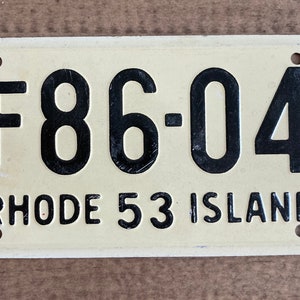 May include: A white metal license plate with black text. The plate reads "F86-04 RHODE 53 ISLAND".