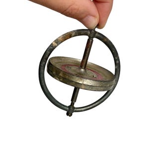 May include: A vintage gyroscope toy. The spinning disc is silver-coloured with a red ring, held within a dark metal frame. The toy is held by a hand against a white background.