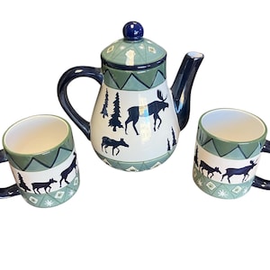 May include: A ceramic teapot and two mugs with a woodland theme. The teapot and mugs are decorated with a green and white background and black silhouettes of moose and trees.
