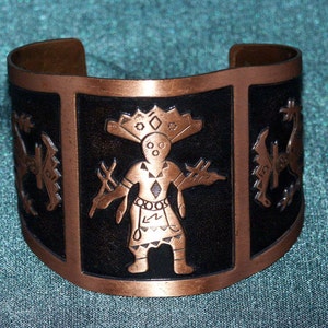 May include: A copper bracelet with a black background and an embossed design of a Native American figure in a headdress. The figure is holding a staff and is surrounded by other symbols.