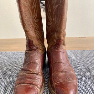 May include: A pair of brown leather cowboy boots with intricate stitching on the shaft. The boots have a pointed toe and a slight heel.