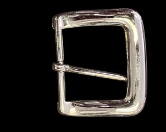 May include: A polished silver metal belt buckle, rectangular in shape with a single bar. The buckle has a classic design and is set against a black background. The metal has a shiny finish.