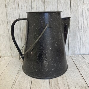 Könnte beinhalten: Schwarzer Emaille-Krug mit gesprenkeltem Finish und einem Drahtgriff. Der Krug hat einen Ausguss und eine breite Öffnung.