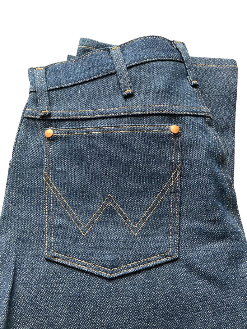May include: Close-up of a pair of blue denim jeans. The jeans feature a classic five-pocket design with a back pocket showcasing a distinctive 'W' stitch pattern. The jeans have copper-colored rivets and tan stitching.
