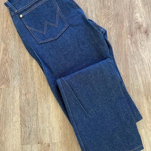 May include: Dark blue denim jeans with a classic five-pocket design. Features a back pocket with a signature stitched design and gold-colored rivets. The jeans are folded, showcasing the straight leg cut and the contrast stitching.