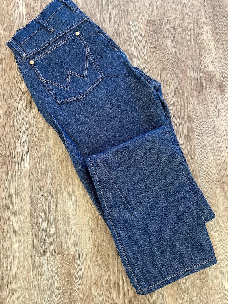 May include: Dark blue denim jeans with a classic five-pocket design. Features a back pocket with a signature stitched design and gold-colored rivets. The jeans are folded, showcasing the straight leg cut and the contrast stitching.
