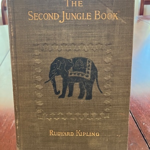 Puede incluir: El Segundo Libro de la Selva de Rudyard Kipling, un libro antiguo de tapa dura con una cubierta de tela verde y una silueta negra de un elefante con una silla de montar decorativa en la portada.