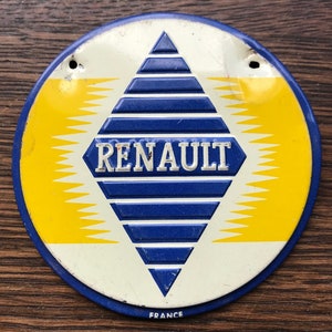 May include: A round, white metal emblem with a blue diamond shape in the center. The diamond has a white outline and is filled with blue and white stripes. The word "RENAULT" is written in white letters on the diamond. The background is yellow with a white border. The words "FRANCE" are written in black at the bottom of the emblem.