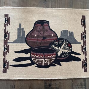 May include: Rectangular beige woven rug with a Southwestern design. The rug features a central image of three pottery vessels in brown, black, and white, with a desert landscape silhouette in the background. The rug has fringed edges.