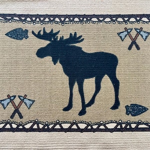 May include: Rectangular tapestry with a tan background and a dark blue moose silhouette in the center. The border is dark brown with a rope-like design. Features crossed axes and arrowheads in each corner, creating a rustic, outdoorsy theme.