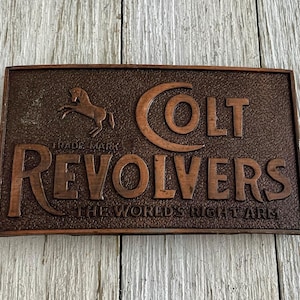 Op de afbeelding: Een rechthoekige, bronskleurige gesp met de woorden "COLT REVOLVERS" in verhoogde letters. Een paardensilhouet staat boven het woord "REVOLVERS". De tekst "TRADE MARK" staat boven het paard en "THE WORLD'S RIGHT ARM" staat onder het woord "REVOLVERS".