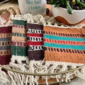 May include: Four woven coasters with fringed edges in a variety of colors, including burgundy, green, and orange. The coasters are arranged on a gold-colored tray, with a candle and a small potted plant in the background.