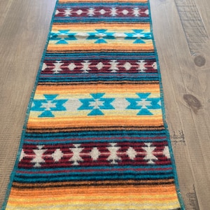 May include: A rectangular, woven table runner with a Southwestern design. The runner features a repeating pattern of turquoise, orange, red, and cream geometric shapes and stripes. The edges are finished with a fringe.