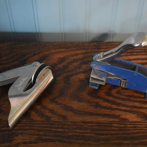 Op de afbeelding: Twee vintage metalen stempels, een zilveren en een blauwe, met handvatten. De zilveren stempel heeft de tekst "Justrite" gegraveerd.