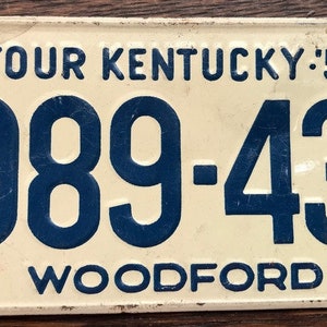 May include: A white metal license plate with blue text. The text reads "Tour Kentucky '54" and "989-431" with the word "Woodford" below the numbers.