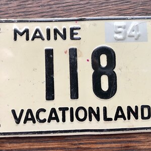 May include: Vintage Maine license plate with the state name at the top and "VACATIONLAND" at the bottom. The plate has the number "118" in black and a sticker with the number "54". The plate is off-white with black lettering.