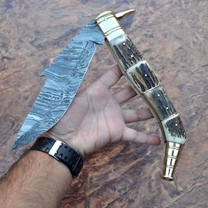 May include: A folding knife featuring a patterned Damascus steel blade and a curved handle made of textured brown stag horn with polished brass bolsters and a decorative brass end cap, showcasing a distinct layered metal aesthetic.