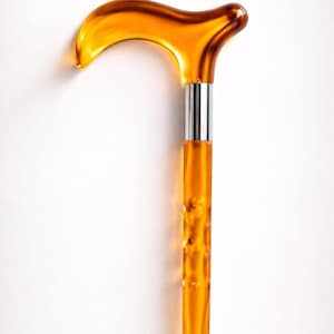 Puede incluir: Bastón de color ámbar con un mango curvo y un eje texturizado. El mango es de un tono ámbar más oscuro, y el eje es de un ámbar más claro y translúcido. Una banda de metal plateado conecta el mango con el eje. El bastón está sobre un fondo blanco.
