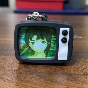 May include: A black retro television-shaped keychain. The screen shows a colourful anime-style image of a girl with green eyes and black hair. The TV has white buttons and a silver chain.